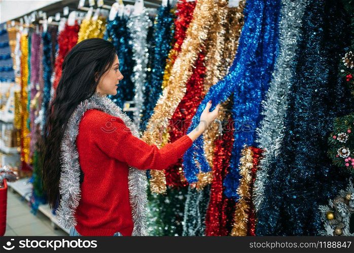 Young woman choosing holiday fluffy garland, purchase of christmas decoration in supermarket, family tradition. December shopping. Woman choosing fluffy garland in supermarket