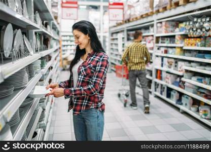 Young woman choosing plates in supermarket. Female customer on shopping in hypermarket, products for the kitchen. Young woman choosing plates in supermarket
