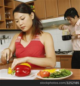 Young woman cutting vegetables in the kitchen with a young man eating noodles in the background