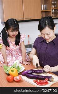 Young woman cutting vegetables with her daughter sitting on kitchen counter