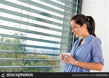Young woman day dreaming looking window blinds drinking tea