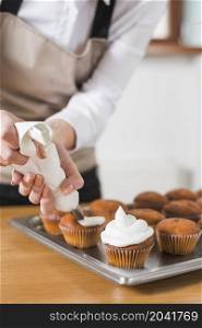 young woman decorating cupcakes with white whipped cream by squeezing confectionery bag
