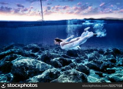 Young woman diving in sunset time, enjoying swimming underwater, wearing long white dress, luxury summer vacation, freedom and pleasure concept