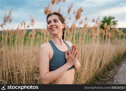 Young woman doing namaste, pranayama yoga breathing exercise on reed natural background. Healthy girl training at summer outdoors. Gratitude, unity with nature, wellness concept. High quality photo. Young woman doing namaste, pranayama yoga breathing exercise on reed natural background. Healthy girl training at summer outdoors. Gratitude, unity with nature, wellness concept.