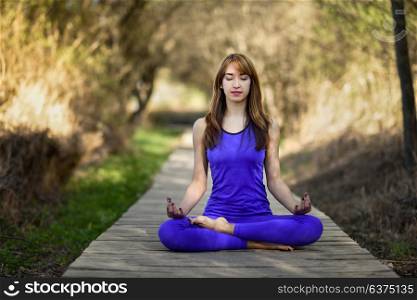Young woman doing yoga in nature. Female wearing sport clothes in lotus figure. Girl on wooden road.