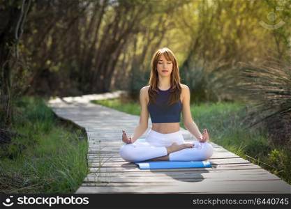 Young woman doing yoga in nature. Female wearing sport clothes in lotus figure. Girl sitting on wooden road.