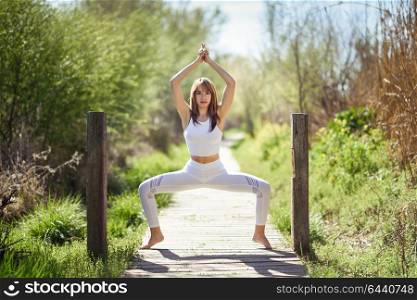 Young woman doing yoga in nature. Female wearing white sport clothes on a wooden road.