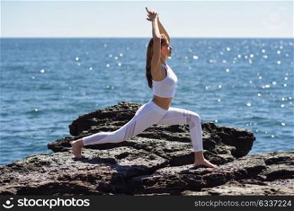 Young woman doing yoga in the beach. Female wearing sport clothes in warrior1 figure with sea at the background.