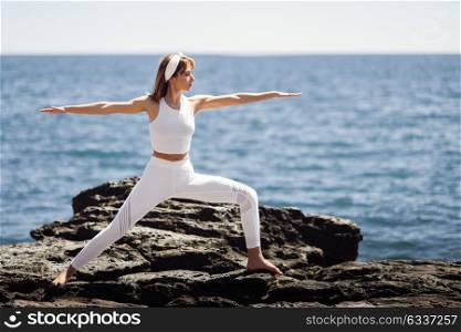 Young woman doing yoga in the beach. Female wearing white sport clothes in warrior 2 figure with defocused sea background.