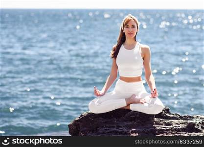 Young woman doing yoga in the beach. Female wearing white sport clothes in lotus figure with defocused sea background.