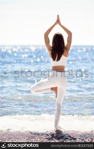 Young woman doing yoga in the beach. Female wearing white sport clothes in tree asana with defocused sea background.