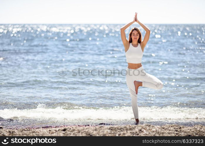 Young woman doing yoga in the beach. Female wearing white sport clothes in tree asana with defocused sea background.