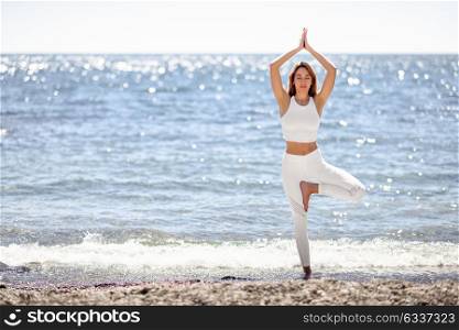 Young woman doing yoga in the beach. Female wearing white sport clothes in tree asana with defocused sea background.