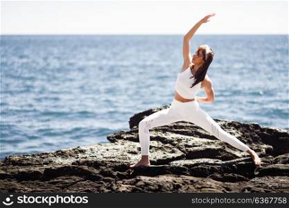 Young woman doing yoga in the beach. Female wearing white sport clothes in warrior at sun figure with defocused sea background.