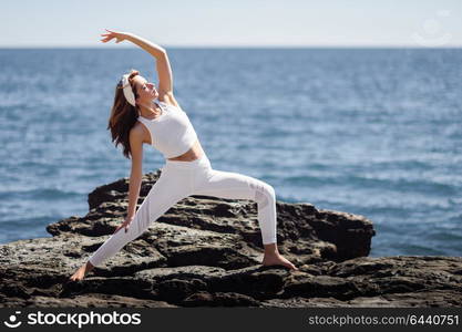 Young woman doing yoga in the beach. Female wearing white sport clothes in warrior at sun figure with defocused sea background.