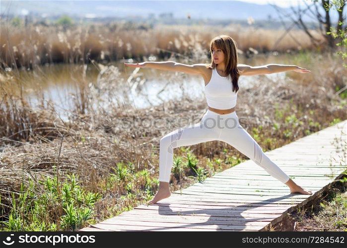 Young woman doing yoga in the beach. Female wearing white sport clothes in warrior 2 figure on wooden road in nature