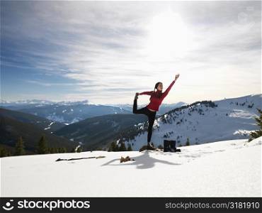 Young woman doing yoga pose on rock in snow in mountainous terrain.