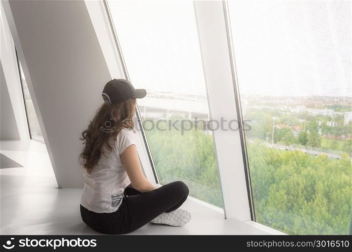 Young woman, dressed casual, sitting directly on the floor and looking out the big windows, towards the beam of light.
