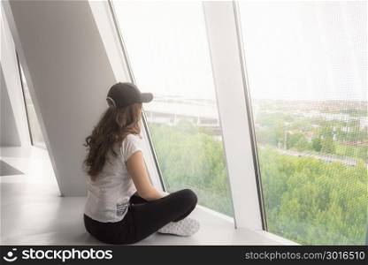 Young woman, dressed casual, sitting directly on the floor and looking out the big windows, towards the beam of light.