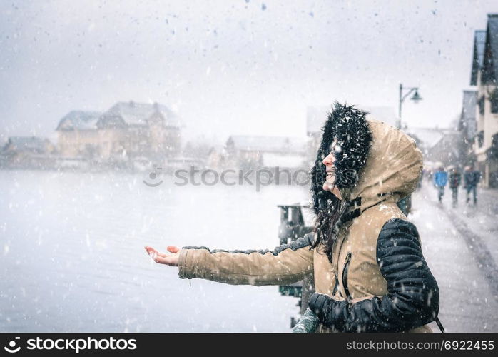 Young woman dressed for cold weather, laughing and holding her hand to catch the snowflakes of the first snowfall in Hallstatt, Austria.