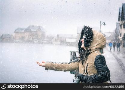 Young woman dressed for cold weather, laughing and holding her hand to catch the snowflakes of the first snowfall in Hallstatt, Austria.