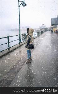 Young woman dressed in a thick jacket for cold weather, standing under snowfall and looking towards the Hallstatter See lake, in Hallstatt, in Austria.