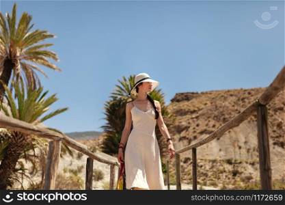 Young woman dressed in a white dress walking on a wooden bridge in the desert with palm trees