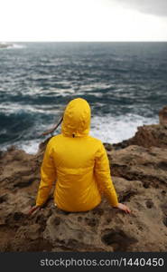 young woman dressed in yellow raincoat sitting on the cliff looking on big waves of the sea while enjoying beautiful sea landscape in rainy day on the rock beach in cloudy spring weather... young woman dressed in yellow raincoat sitting on the cliff looking on big waves of the sea while enjoying beautiful sea landscape in rainy day on the rock beach in cloudy spring weather