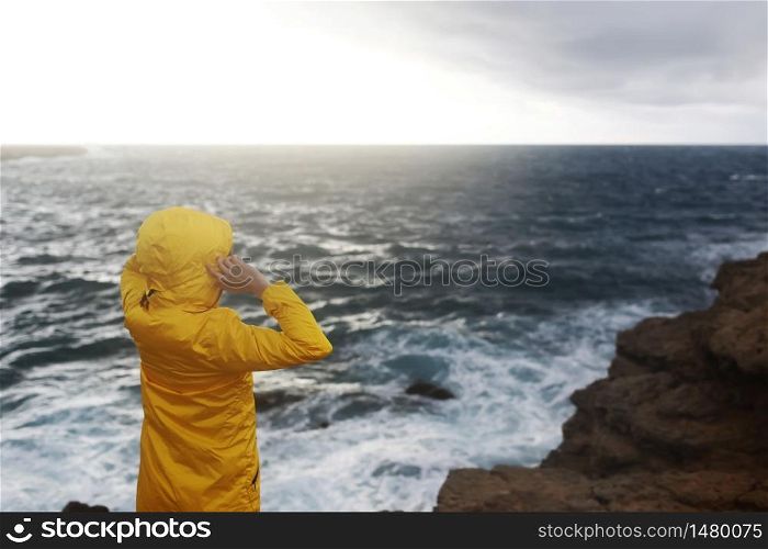 young woman dressed in yellow raincoat standing on the cliff looking on big waves of the sea while enjoying beautiful sea landscape in rainy day on the rock beach in cloudy spring weather.. young woman dressed in yellow raincoat standing on the cliff looking on big waves of the sea while enjoying beautiful sea landscape in rainy day on the rock beach in cloudy spring weather