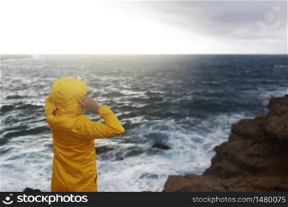 young woman dressed in yellow raincoat standing on the cliff looking on big waves of the sea while enjoying beautiful sea landscape in rainy day on the rock beach in cloudy spring weather.. young woman dressed in yellow raincoat standing on the cliff looking on big waves of the sea while enjoying beautiful sea landscape in rainy day on the rock beach in cloudy spring weather