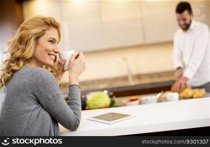 Young woman drinking coffee at the kitchen table while man preparing food