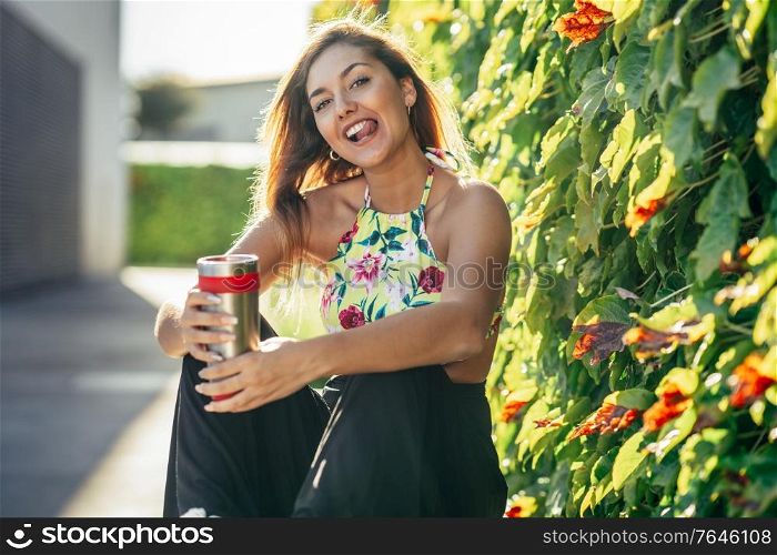 Young woman drinking from a metal water bottle wearing casual clothes. Girl sitting outdoors.. Girl drinking from a metal water bottle wearing casual clothes