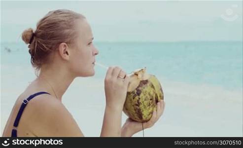 Young woman drinking from coconut sitting by the sea on the beach. She enjoying the view