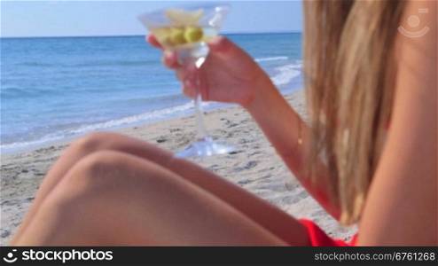 Young woman drinking martini cocktail on the summer sandy beach, focus on background