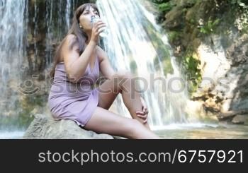 young woman drinking water while sitting near the waterfall