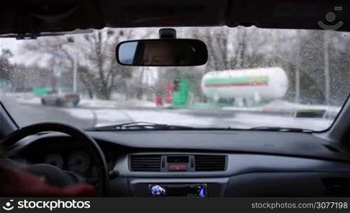 Young woman driver driving car to fuel at gas station in wintertime. View from inside of car. Beautiful girl driving her automobile on winter road heading to petrol station.