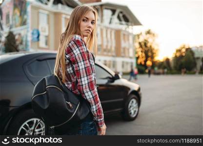 Young woman driver with travel bag against a car. Female person and vehicle, automobile driving concept. Young woman driver with bag against a car