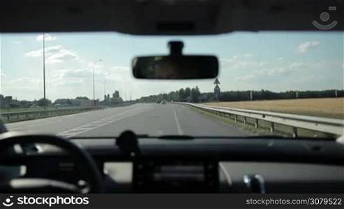 Young woman driving car on highway, passing by crossroad with road signs on background. Female travelling by car during summer vacation trip. View from inside of vehicle.