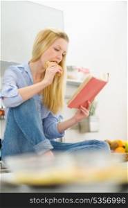Young woman eating sandwich and reading book in kitchen