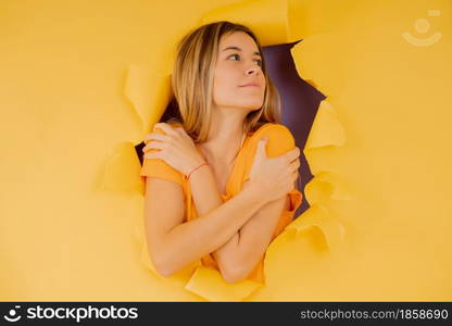 Young woman embracing herself while stands through a hole on the paper wall.