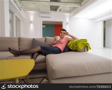 young woman enjoying free time drinking tea while relaxing on corner sofa at big stylish duplex apartment