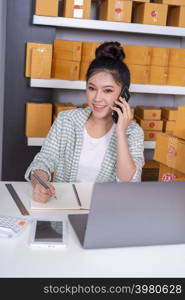 young woman entrepreneur talking with customer on mobile phone and writing product order at home office