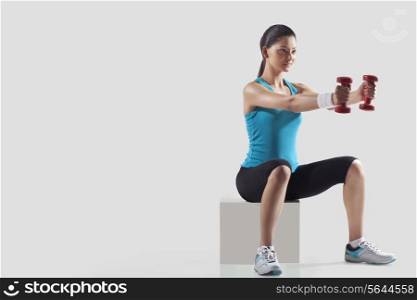Young woman exercising with dumbbells while sitting on stool isolated over gray background