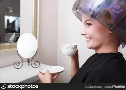 Young woman female client drinking hot drink coffee tea in hairdressing beauty salon. Girl in hair rollers curlers with hairdryer dryer relaxing by hairdresser hairstylist.