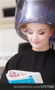 Young woman female client reading magazine in hairdressing beauty salon. Girl in hair rollers curlers with hairdryer dryer relaxing by hairdresser hairstylist. Indoor