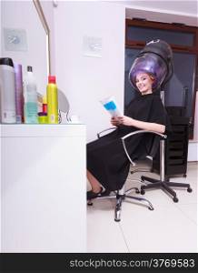 Young woman female client reading magazine in hairdressing beauty salon. Girl in hair rollers curlers with hairdryer dryer relaxing by hairdresser hairstylist.