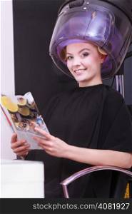 Young woman female client reading magazine in hairdressing beauty salon. Girl in hair rollers curlers with hairdryer dryer relaxing by hairdresser hairstylist.