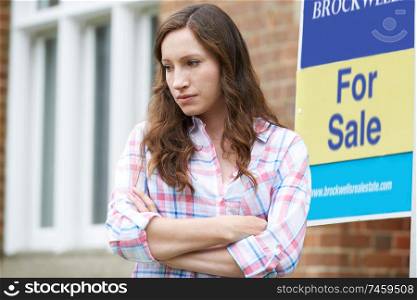 Young Woman Forced To Sell Home Through Financial Problems Standing Outdoors Next To For Sale Sign