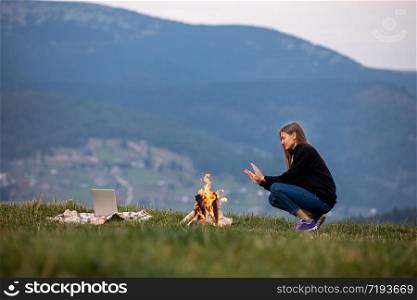 young woman freelancer sits by the fire and warms his hands in the mountains in the evening. tourist girl resting and working on laptop outdoor.. young woman freelancer sits by the fire and warms his hands in the mountains in the evening. tourist girl resting and working on laptop outdoor