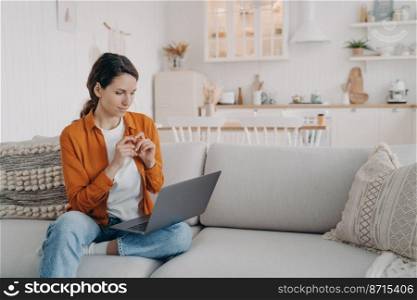Young woman freelancer using laptop working on project sitting on comfortable couch at home. Beautiful female chatting on social networks, shopping online, watching movie or series on sofa.. Female working at laptop, sitting on sofa, chatting on social networks, shopping online. E commerce